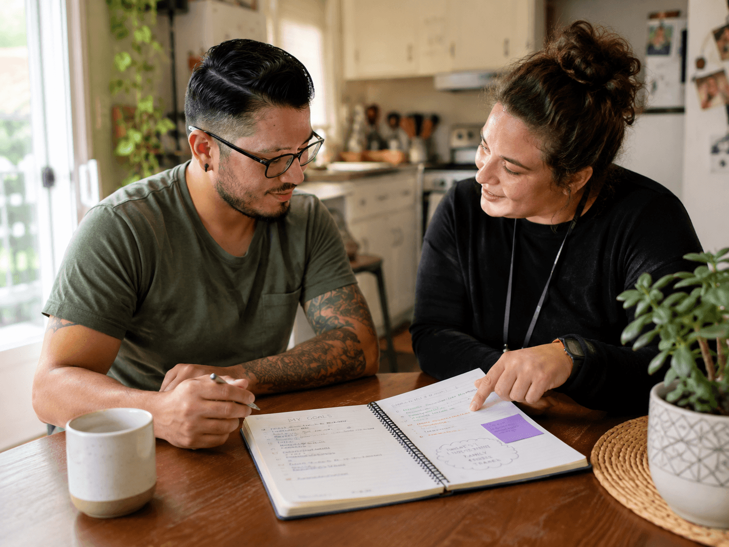 A person reviews personal goals and routines in a planning notebook with their support coordinator at the kitchen table.