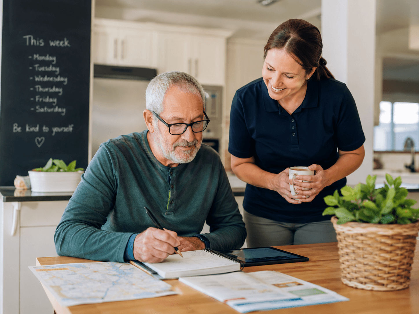 An older adult writes in his weekly planner at the kitchen table while a support professional looks on, smiling, coffee in hand.